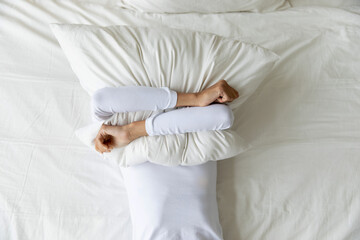 Top view depressed woman covering face with pillow, lying on bed at home alone, frustrated unhappy...
