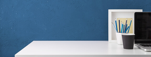 Home office desk with blue wall and white table with coffee cup, laptop, office supplies,  office objects and laptop.