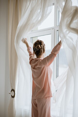 Красивая женщина в розовой пижаме открывает шторы и смотрит в окно