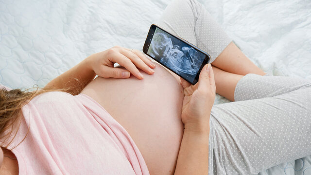 Young pregnant woman in pajamas browsing ultrasound image of her unborn baby. Concept of expecting baby, pregnancy and healthcare