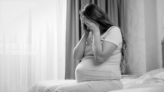 Black and white image of young pregnant woman suffering from depression crying on bed. Concept of maternal and pregnancy depression