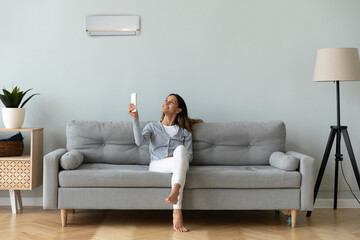 Smiling woman using air conditioner remote controller, sitting on cozy couch at home full length,...