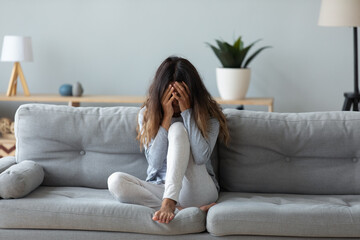 Obraz premium Frustrated woman covering face and crying, sitting on couch at home alone, upset young female suffering from break up with boyfriend or divorce, bad relationship, feeling lonely and depressed