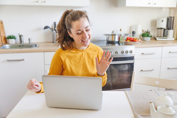 Smiling girl waving hand video calling family by webcam. Woman with laptop having virtual meeting chat video call conference sitting on kitchen at home. New normal social distance self isolation.