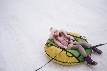 Fototapeta premium Little cute girl in pink warm outwear having fun rides inflatable snow tube in snowy white cold winter outdoors. Family sport vacation activities.