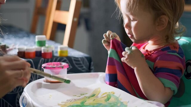 Baby and paints. Toddler girl licks her paint stained shirt and enjoys art with her mother at home