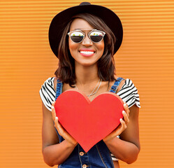 Beautiful smiling african woman holding a red heart. Fashion portrait stylish pretty woman in sunglasses outdoor.