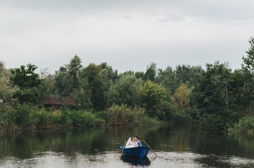 Stylish groom in a suit and a cute bride in a white lace dress are sitting in a wooden boat, walking and swimming on the lake, enjoying the beautiful nature. Wedding portrait.