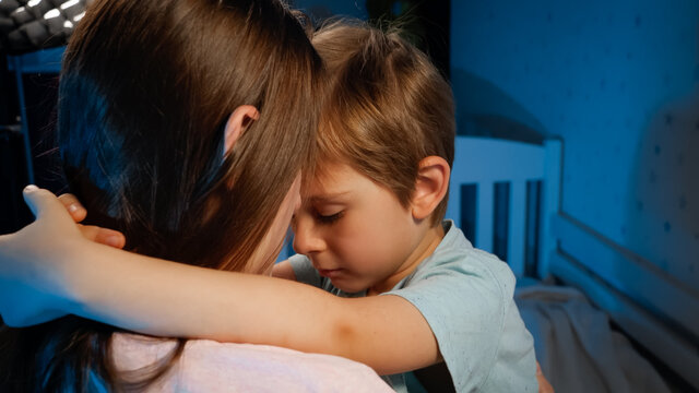Portrait of little upset and sad toddler boy hugging and kissing his mother before going to sleep at night. Loving and caring parents and children