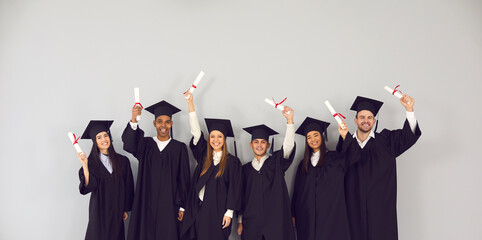 Fototapeta Study abroad website banner. Group of happy smiling diverse academy graduates holding up diplomas. International university students in traditional black academic gowns and caps celebrating graduation obraz