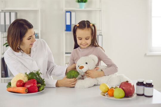 Doctor Teaching Child To Eat Fresh Healthy Food. Dietitian Or Nutritionist Talking To Little Girl, Playing Fun Game Of Feeding Teddy Bear And Telling About Benefits Of Eating Raw Fruit And Vegetables