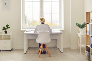 Back view of woman busy with paperwork, sitting at table by the window in hospital or clinic...