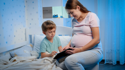 Young smiling pregnant mother sitting next to her child lying in bed and watching video on tablet computer