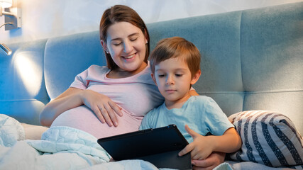 Happy smiling boy with mother watching cartoons on tablet computer in bed at night