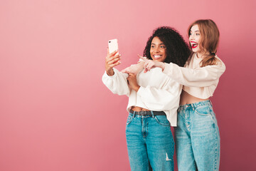 Two young beautiful smiling international hipster female in trendy summer jeans clothes. Sexy carefree women posing near pink wall in studio. Positive models having fun. They taking selfie photos
