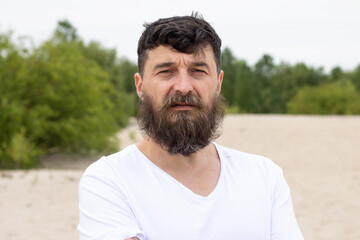 Portrait of a serious bearded man in a white shirt looking at the camera against the background of nature.