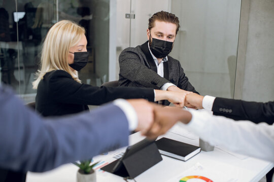 Close up cropped image of happy business coworkers with face protective masks, bumping their fists after successful meeting in the office. Focus on blond lady bumping fist with male colleagues