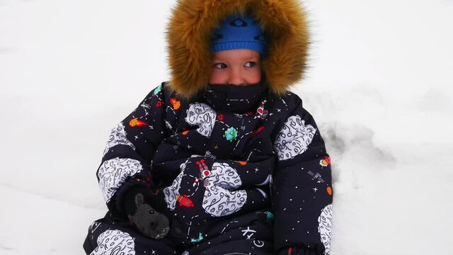 A little boy in a down jacket sits in a snowdrift close-up