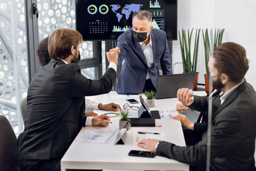 Confident mature male director in facial protective mask, friendly greeting with fist bump every member of his multiethnic team, during business meeting at the time of coronavirus pandemic