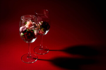 Flowers lie in wine glasses on a red background. Roses and peonies in a glass. The shadow from the glasses on the red surface. Free space. Romance. Festive decoration.