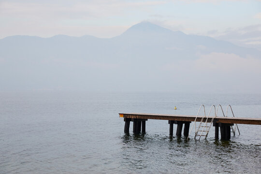 Pontile di legno vuoto su Lago di Garda con scaletta e montagne sullo sfondo