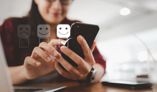 Customer service evaluation concept. smiling Asian female Is using a smartphone And she is pressing face emoticon smiling in satisfaction on virtual touch screen.