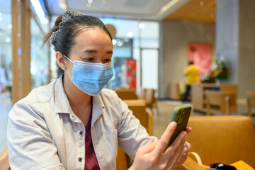 New normal of Asian lady wearing surgical face mask using mobile phone at coffee shop or cafe. Ways of life after the coronavirus COVID-19 outbreak , watch and protect yourself in public places.