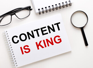 CONTENT IS KING is written in a white notebook on a light background near the notebook, black-framed glasses and a magnifying glass.