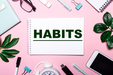 The word HABITS is written in a white notebook on a pink background surrounded by business accessories and green leaves.