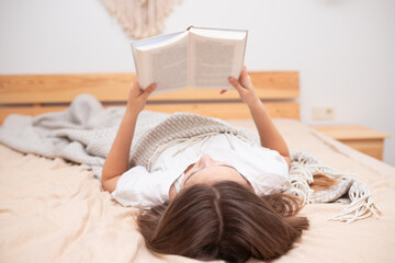 Teenage girl lying on the bed with a blanket and reading a book. Home schooling. Self-isolation during quarantine. Stay home. Social distancing. Coronavirus