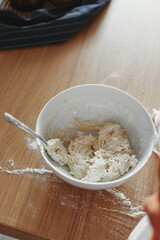 White bowl with dough and spoon in it, on the wooden table in the kitchen. The process of mixing ingredients for pastry. Closeup, selective focus.