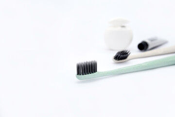 A pastel green toothbrush with thin black bristles in the foreground, and a beige toothbrush, white dental floss and a travel tube of toothpaste in the background on a white background with copy space