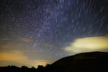 A trail of bright stars from the Milky Way in the cloudy night sky over the hills.
