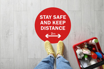 Woman near sign STAY SAFE AND KEEP DISTANCE in supermarket, top view. Coronavirus pandemic