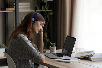 Exhausted millennial female student in earphones sit at desk overwhelmed with study or work online on laptop. Tired young Caucasian woman suffer from headache or migraine from computer job.