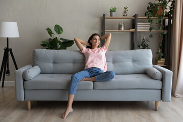Obraz premium Happy millennial indian Arabic woman sit rest on comfortable couch in modern living room look in distance dreaming. Smiling young ethnic female renter or tenant relax on sofa at home, enjoy weekend.