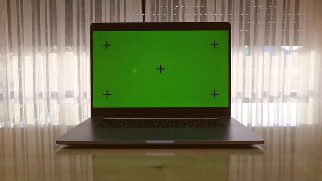 Laptop with the green screen on the table and window opens on the background. Sun shining outside the room.