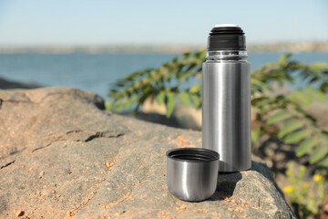 Modern silver thermos on stone near river. Space for text