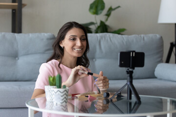Smiling young indian Arabic female vlogger record makeup tutorial on smartphone camera at home. Happy millennial ethnic woman coach or influencer shoot cosmetics haul or unpacking on cellphone.
