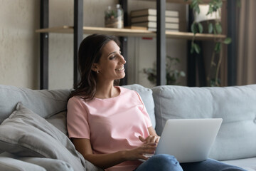 Happy millennial indian Arabic woman relax on sofa at home use laptop look in distance dreaming. Smiling young middle eastern female rest on couch with computer visualize or imagine opportunities.