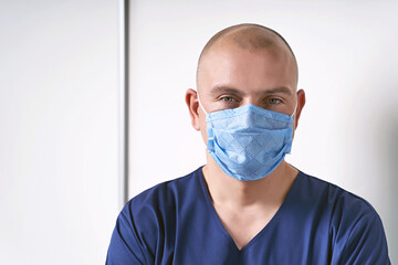 Experienced male massage therapist masseur in his medical office standing by a massage table wearing professional uniform and disposable face mask.