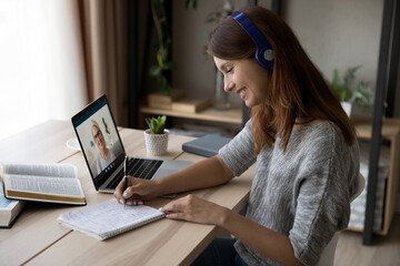 Smiling millennial female student in earphones make notes have online webcam lesson with teacher or tutor on computer. Happy young woman study on internet, talk on video call with coach on laptop. - obrazy, fototapety, plakaty