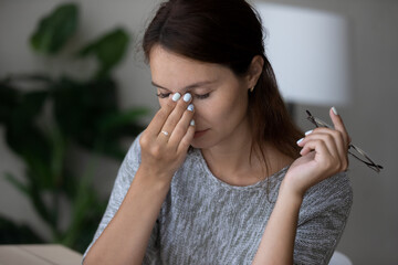 Close up of tired millennial female take off glasses massage eyes suffering from headache or migraine. Exhausted unwell young woman struggle with blurry vision or dizziness from computer work.