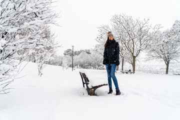Fototapeta na wymiar Chica joven en un parque nevado en un día de invierno con gorro de lana gris, jeans y cazadora negra