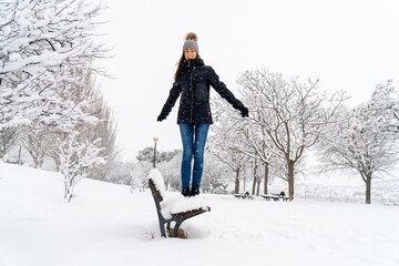 Chica joven en un parque nevado en un día de invierno con gorro de lana gris, jeans y cazadora...