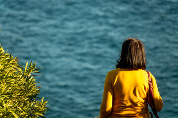 Woman from behind with yellow sweater next to a green plant looking at the blue sea