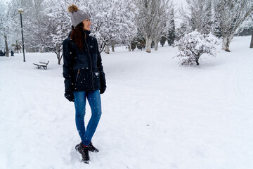 Chica joven en un parque nevado en un día de invierno con gorro de lana gris, jeans y cazadora negra