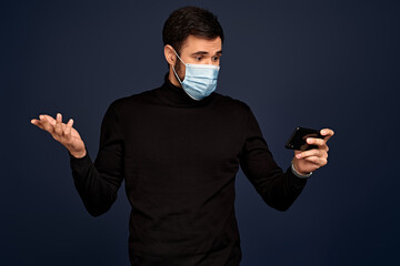 Upset young man in black sweater, with sterile face mask making video call with mobile phone, spreading hand isolated on Pacific Blue background. Coronavirus 2019-ncov sars covid-19 flu virus concept.