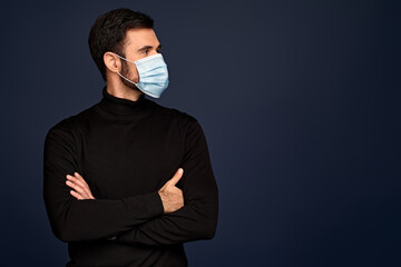 Young man in black sweater, sterile face mask looking aside holding hands folded isolated on...