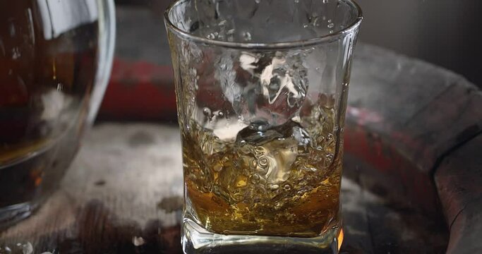 An ice cube slowly falls into a glass of whiskey on top of a vintage whiskey barrel. A decanter with a drink stands nearby, a dark brown background. Blackmagic Ursa Pro G2. 